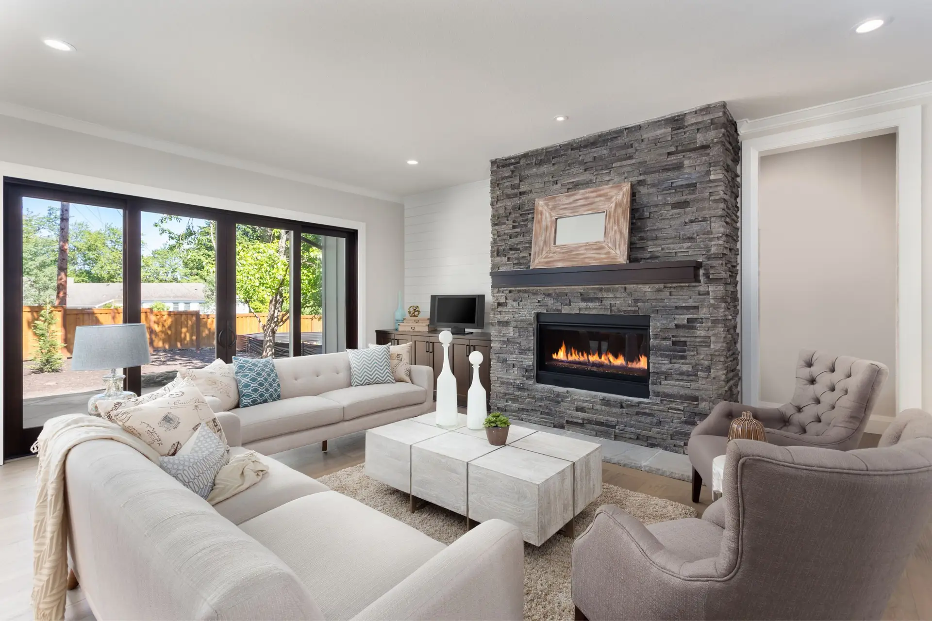 Modern living room with a stone fireplace, beige sofas, and armchairs. Large windows provide natural light, showcasing a backyard. A flat-screen TV is mounted above the fireplace, perfect for an Airbnb agency seeking to enhance guest experiences in short-term rental properties.