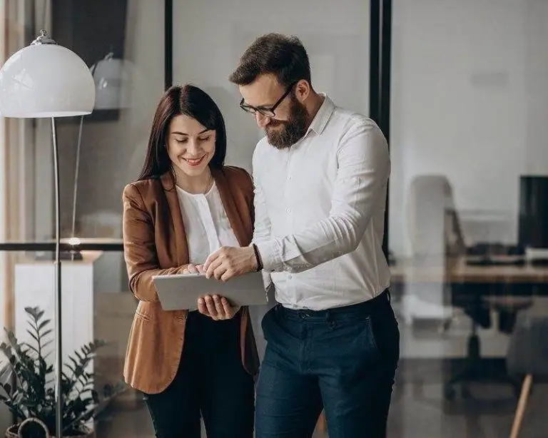 Two people stand in an office, looking at a tablet together. One is holding the tablet, and the other is pointing at it. There is a lamp and a plant in the background.