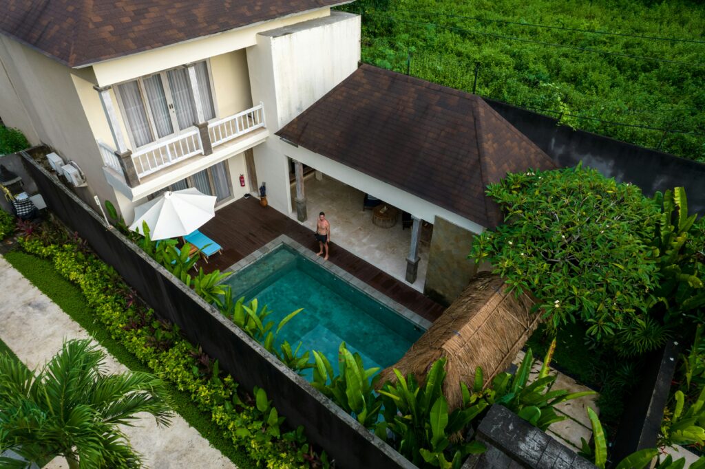 Aerial view of a person standing near a rectangular pool in a backyard, surrounded by greenery, with a two-story house featuring a balcony and a thatched pavilion.