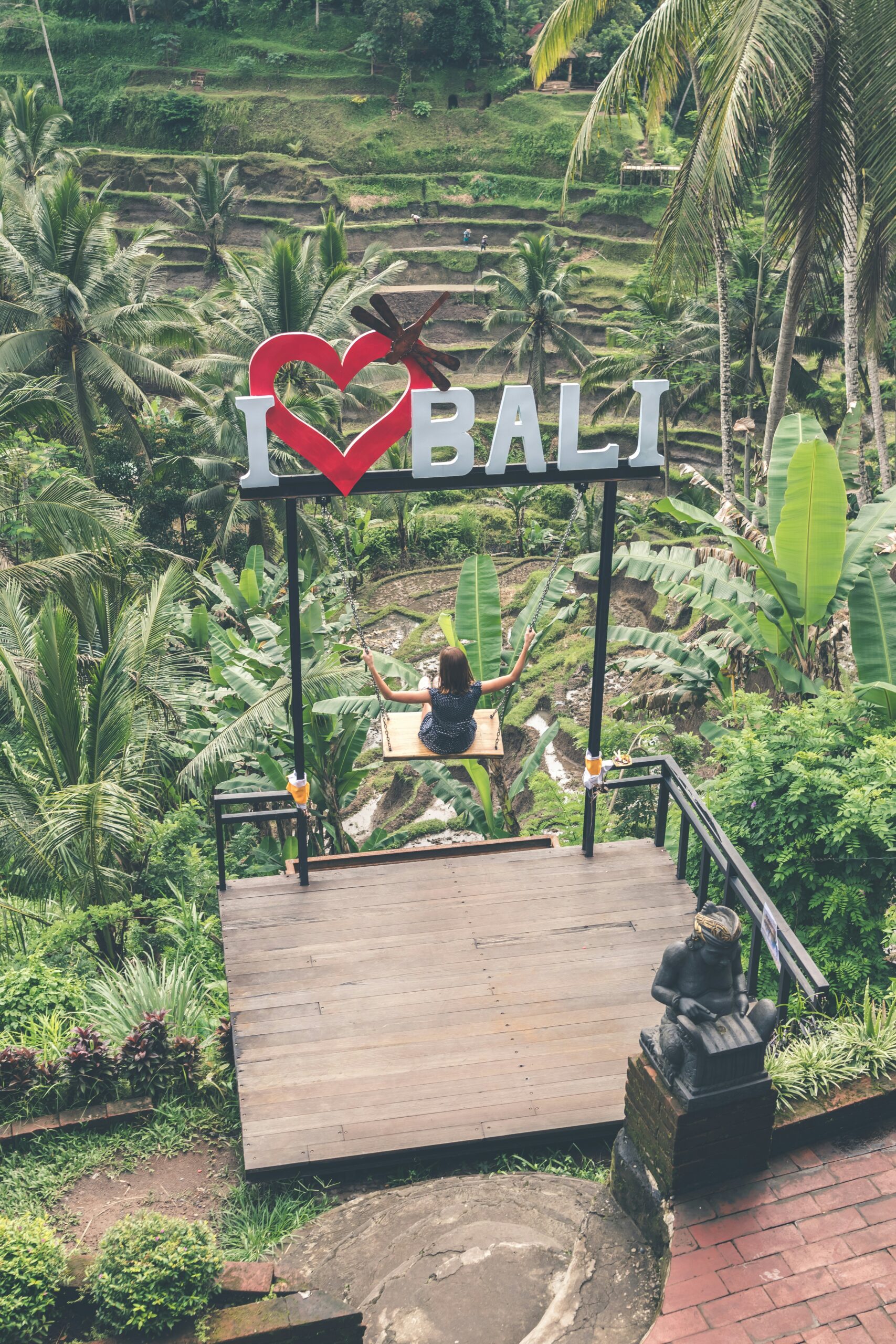 Person sitting on a swing with "I ♥️ BALI" sign above, overlooking lush rice terraces and palm trees in Bali.