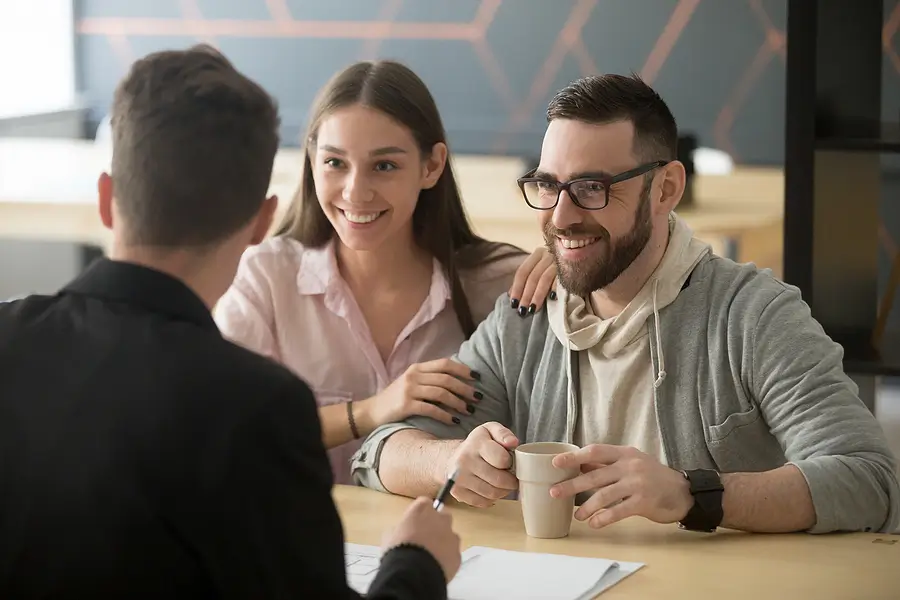 Three people sit at a table in a business setting. A man and woman smile while speaking with a person sitting across from them. The man holds a mug. Papers are visible on the table.