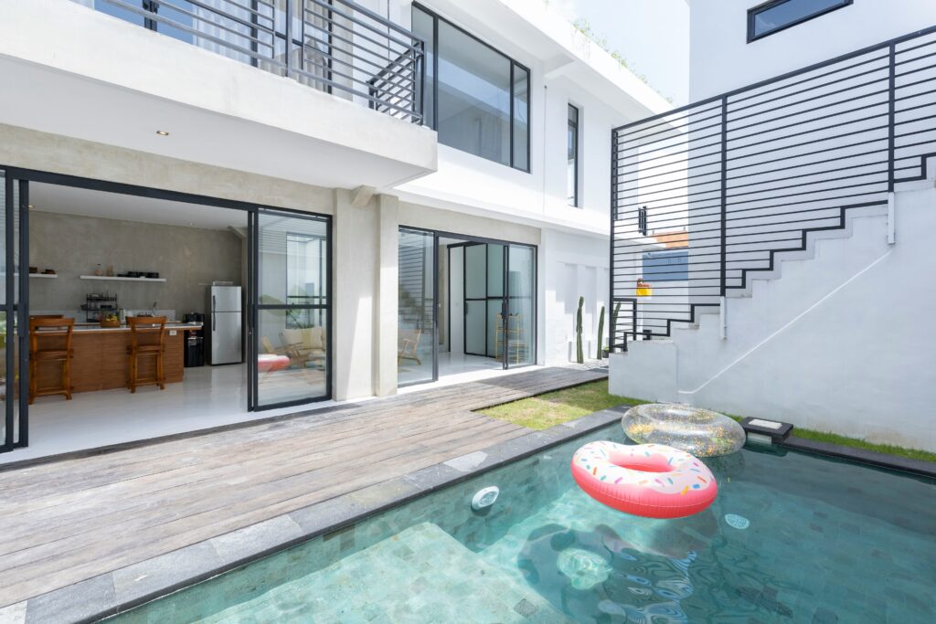 Modern house with sliding glass doors opening to a wooden deck and small pool. Inflatable toys float in the water. Indoor view shows kitchen and dining area. Stairs lead to upper level.