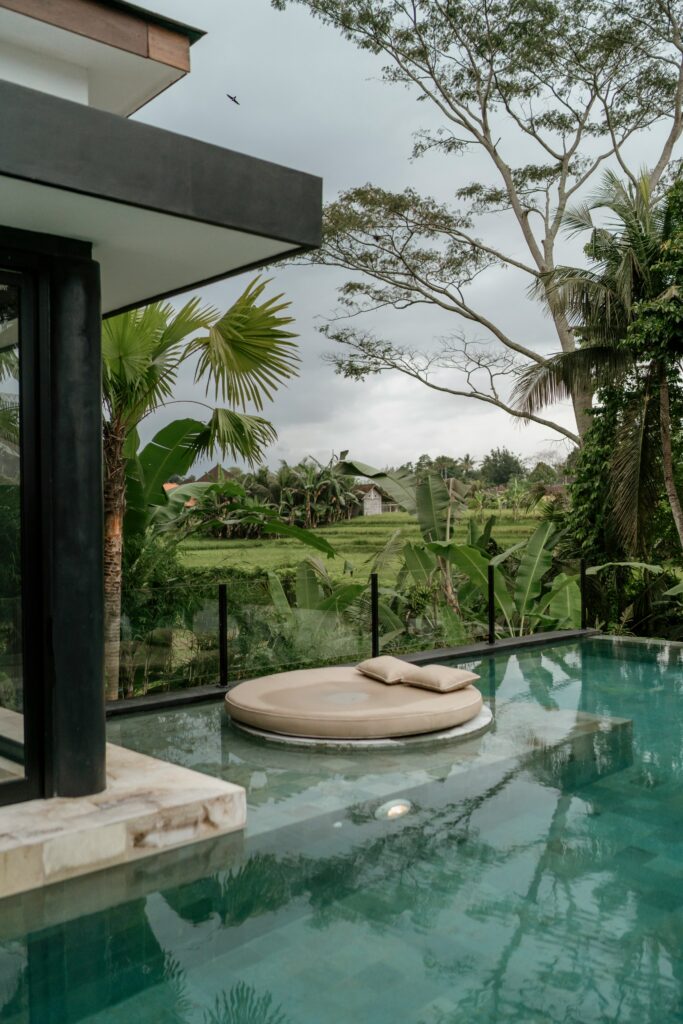 Modern villa with a pool featuring a floating round lounge chair, surrounded by lush tropical greenery and trees under a cloudy sky.