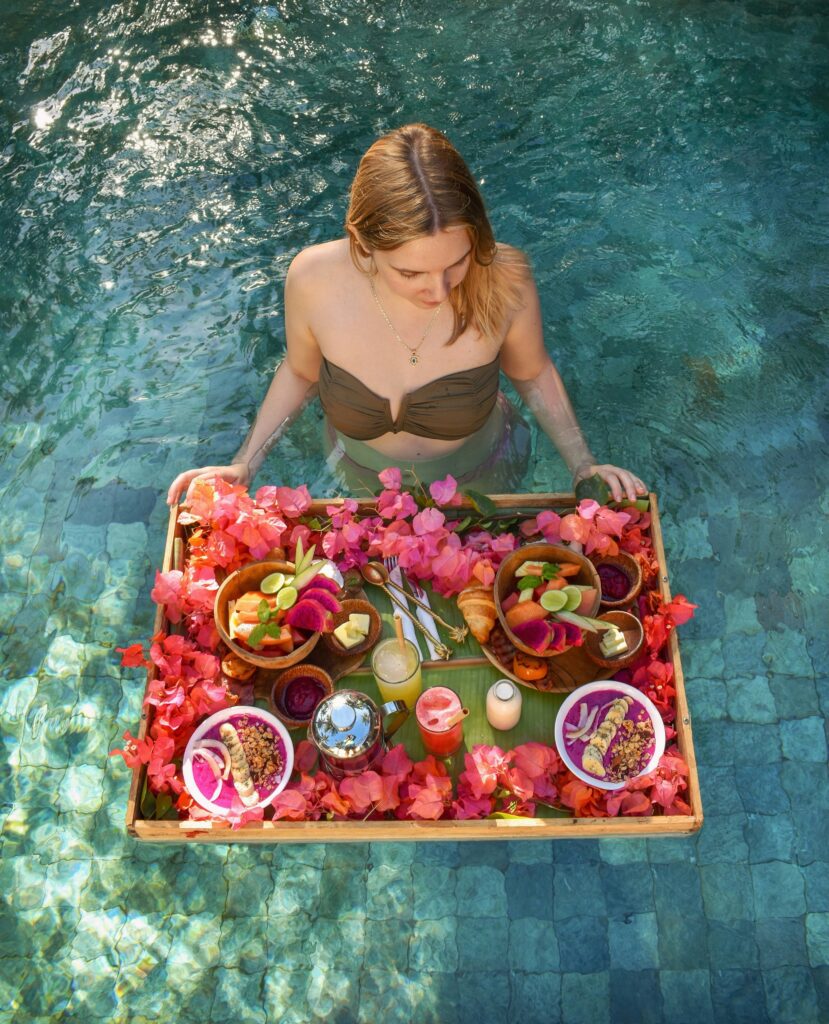 A woman in a pool holds a floating tray with a variety of foods and drinks, surrounded by pink flowers. She is wearing a green swimsuit.