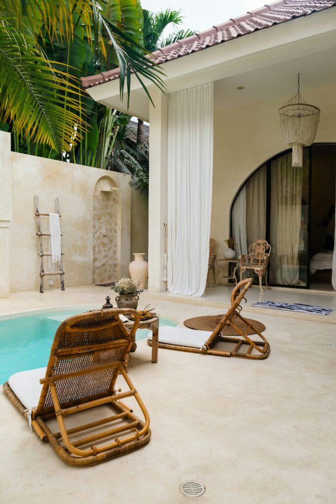 Tropical poolside scene with two wooden lounge chairs, a small pool, and a partially open door leading to an indoor space. Lush greenery is visible around the area.