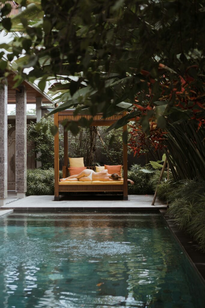 Luxurious outdoor daybed with orange pillows next to a turquoise pool, surrounded by lush greenery and tropical plants.