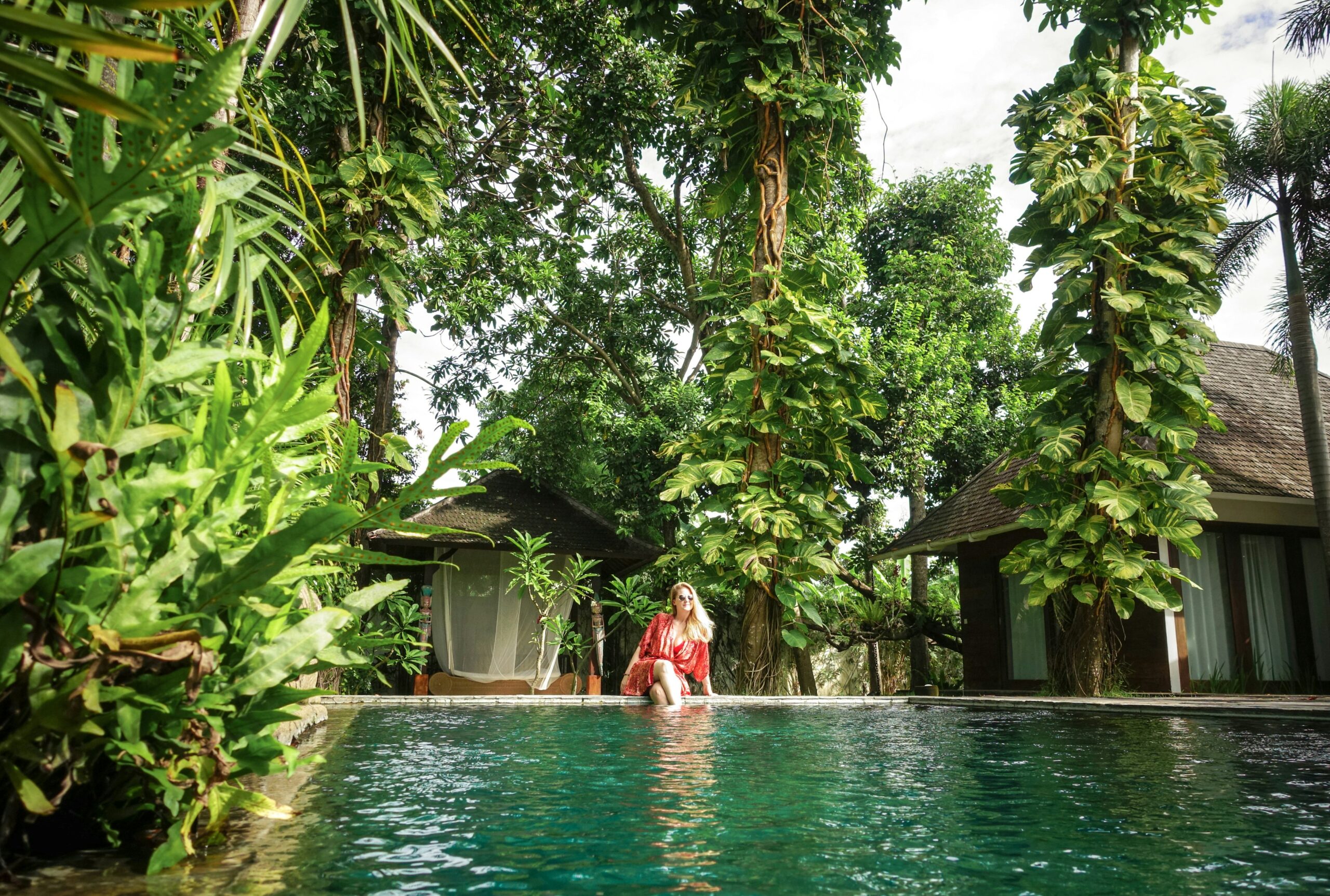 Person in red sitting by a pool, surrounded by lush greenery and two huts in the background.