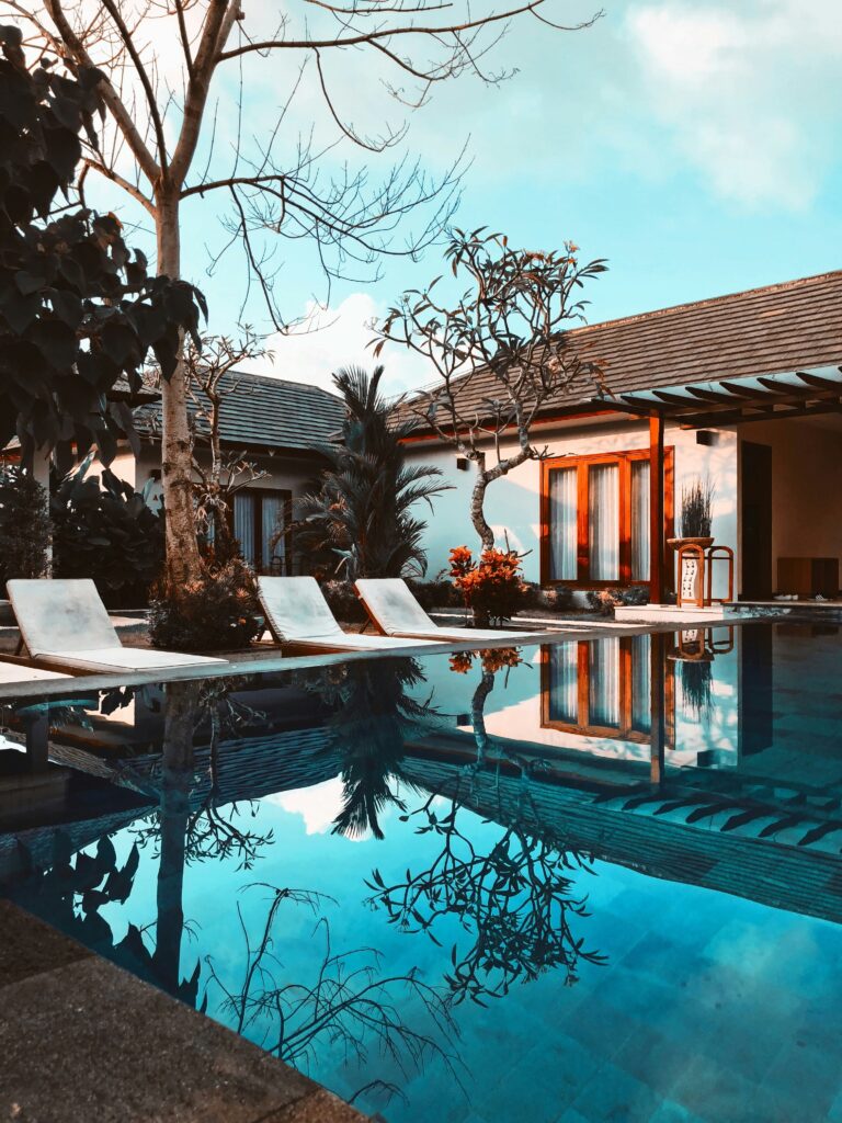 A serene backyard pool with four lounge chairs, surrounded by trees and a modern house. The clear water reflects the sky and branches, creating a tranquil setting.