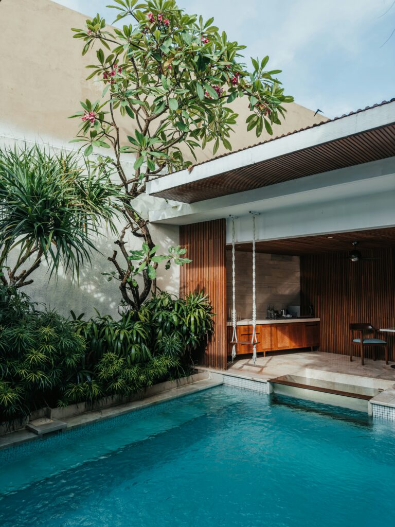 Outdoor pool area with a wooden swing and lush greenery, including trees and shrubs. An open wooden kitchen area is visible in the background.