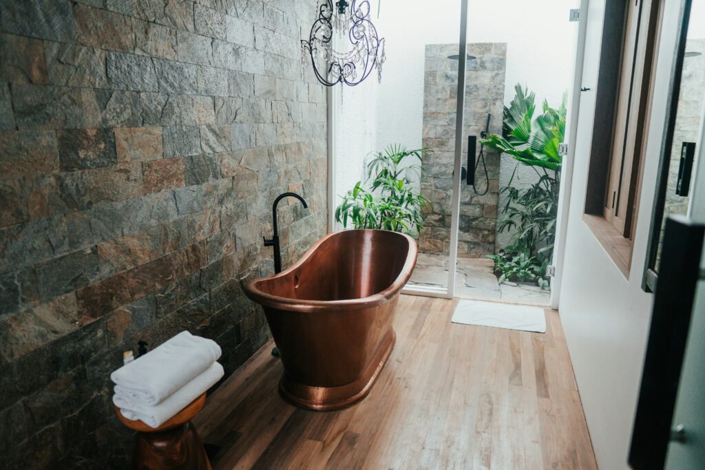 Bathroom with a copper bathtub, stone accent wall, wooden floor, and a glass shower door. Towels are neatly folded on a small stool beside the tub. Green plants are outside the shower area.